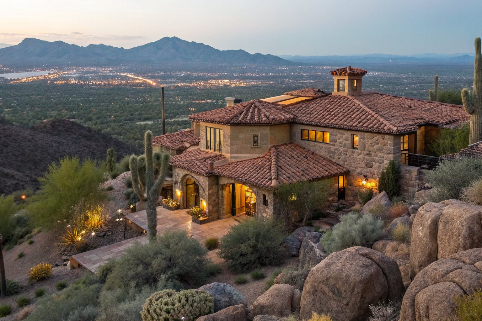 A large stone and stucco home with a clay tile roof on a rocky hillside among saguaros at dusk, city lights below