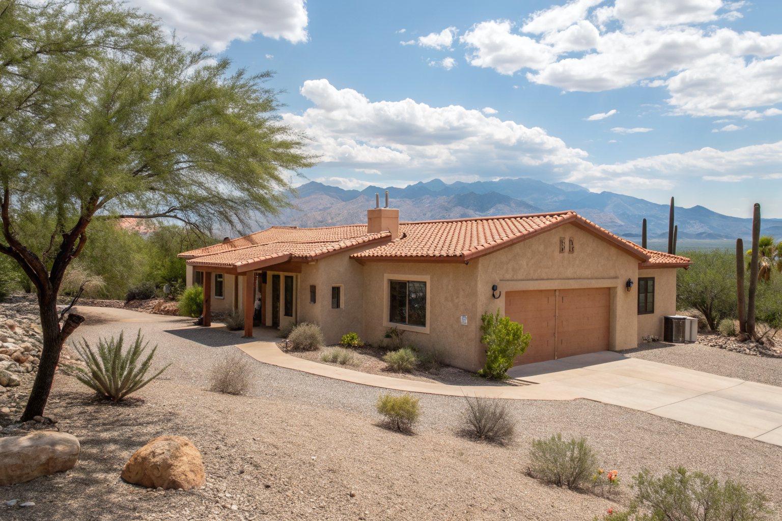 A single-story tan stucco home with a terracotta tile roof, a palo verde tree and saguaros, mountains behind